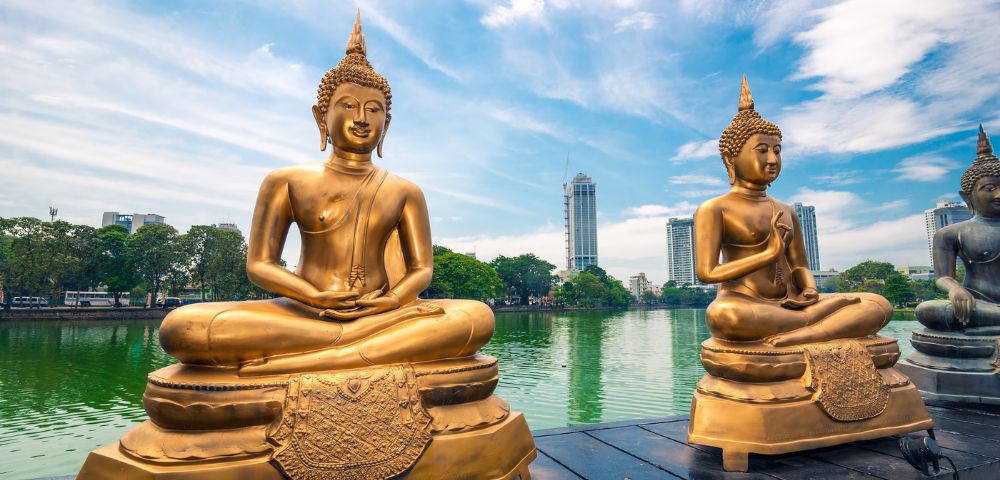 Two golden Buddha statues sit serenely by a tranquil lake, set against a backdrop of lush trees and modern skyscrapers under a partly cloudy sky.