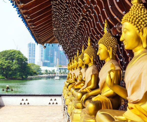 Golden Buddha statues sit in serene, meditative poses under a wooden pavilion overlooking a tranquil lake, with city skyscrapers in the background.
