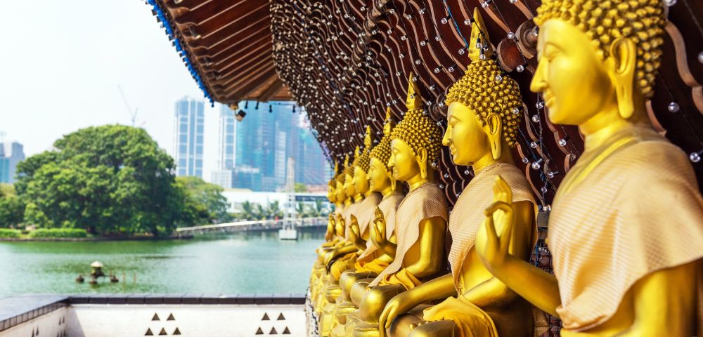 Golden Buddha statues sit in serene, meditative poses under a wooden pavilion overlooking a tranquil lake, with city skyscrapers in the background.