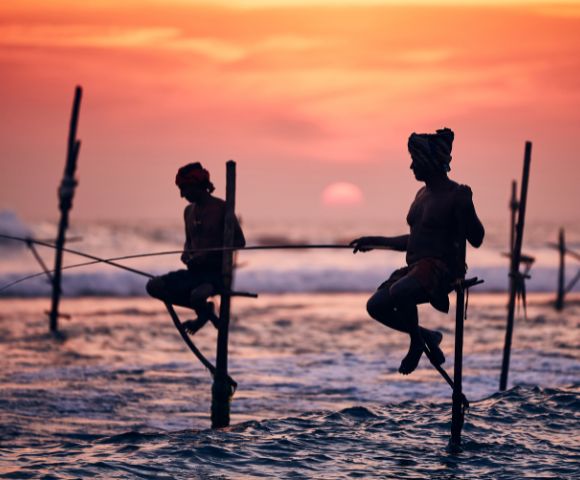 Two fishermen sit on stilts above the ocean at sunset, casting lines into the waves. The sky glows orange, creating a serene and timeless scene.