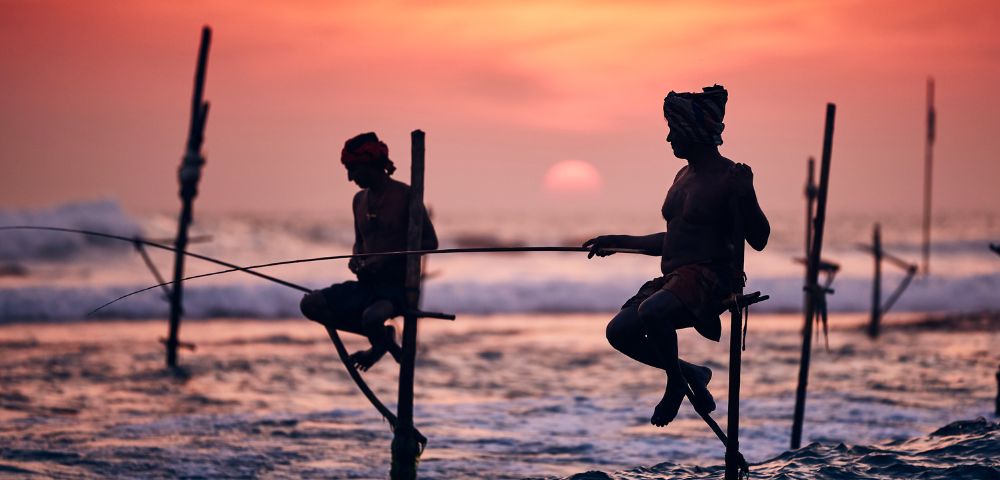 Two fishermen sit on stilts above the ocean at sunset, casting lines into the waves. The sky glows orange, creating a serene and timeless scene.