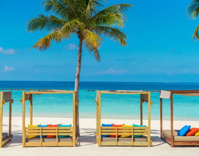Tropical beach scene with wooden cabanas, colorful cushions, palm trees, and turquoise ocean under a clear blue sky. Relaxing and inviting atmosphere.