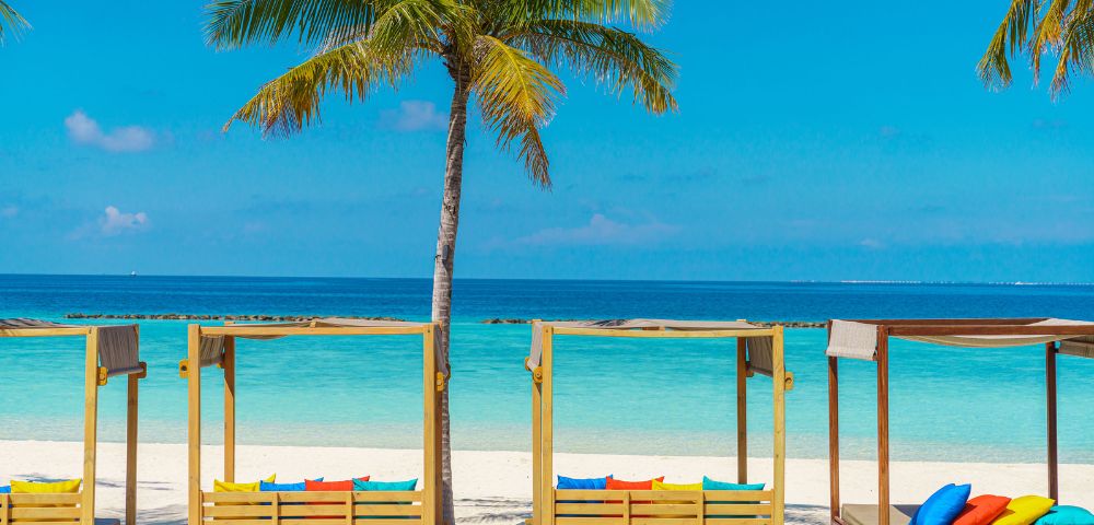 Tropical beach scene with wooden cabanas, colorful cushions, palm trees, and turquoise ocean under a clear blue sky. Relaxing and inviting atmosphere.