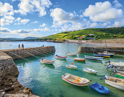 A serene coastal scene with colorful boats floating in a calm harbor. Green hills and cloudy blue sky in the background; two people walk on a pier.