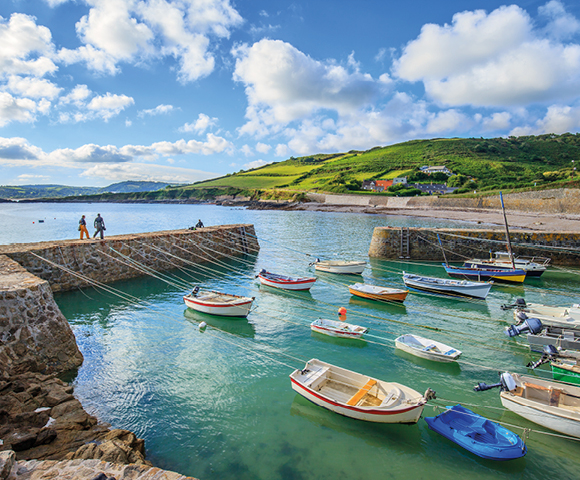A serene coastal scene with colorful boats floating in a calm harbor. Green hills and cloudy blue sky in the background; two people walk on a pier.