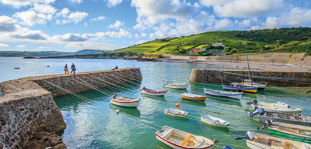 A serene coastal scene with colorful boats floating in a calm harbor. Green hills and cloudy blue sky in the background; two people walk on a pier.