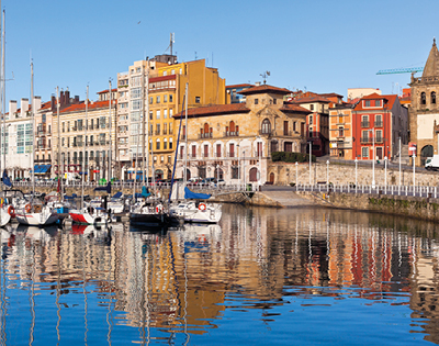 A serene harbor scene with colorful buildings lining the waterfront. Boats are docked on calm, reflective water under a clear blue sky.