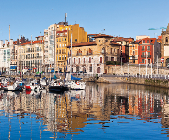 A serene harbor scene with colorful buildings lining the waterfront. Boats are docked on calm, reflective water under a clear blue sky.