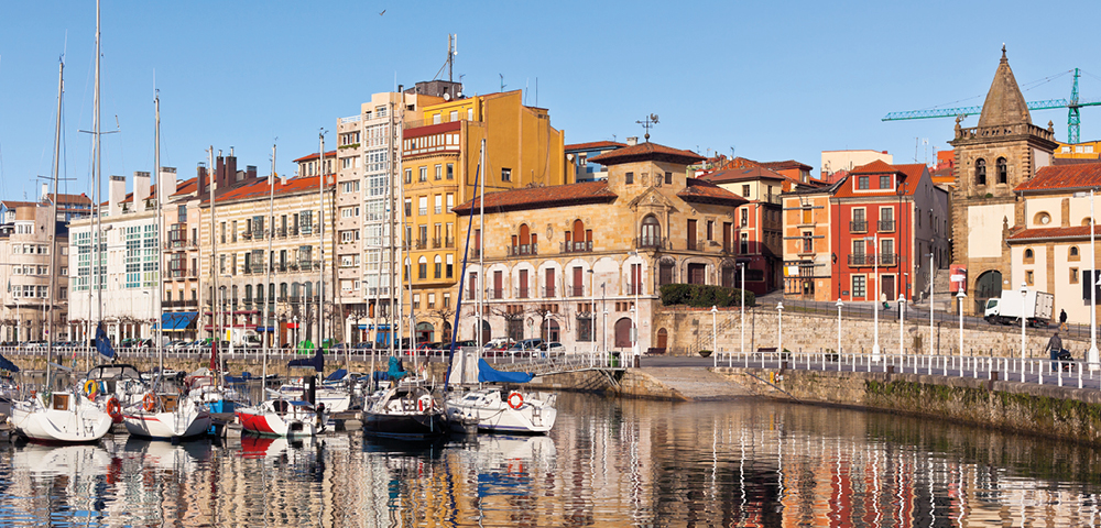 A serene harbor scene with colorful buildings lining the waterfront. Boats are docked on calm, reflective water under a clear blue sky.