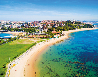 A coastal cityscape featuring a sandy beach curving along turquoise water, with lush green parkland in the foreground and urban buildings on a hill.
