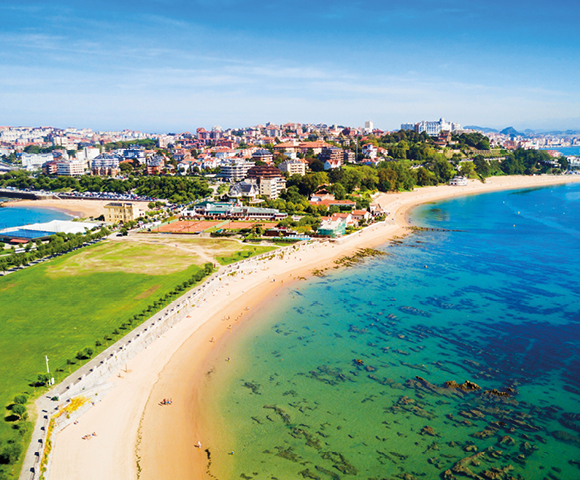 A coastal cityscape featuring a sandy beach curving along turquoise water, with lush green parkland in the foreground and urban buildings on a hill.