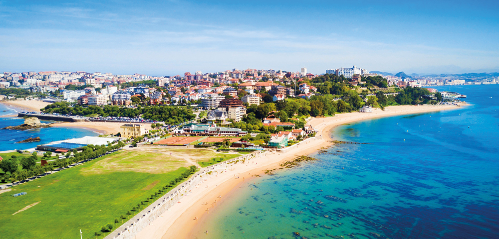A coastal cityscape featuring a sandy beach curving along turquoise water, with lush green parkland in the foreground and urban buildings on a hill.