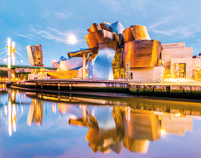 Modern, metallic museum building with curved, angular shapes reflecting in a calm river during twilight. A nearby bridge is illuminated, creating a vibrant cityscape.
