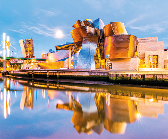 Modern, metallic museum building with curved, angular shapes reflecting in a calm river during twilight. A nearby bridge is illuminated, creating a vibrant cityscape.