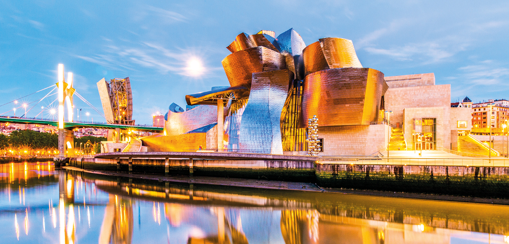 Modern, metallic museum building with curved, angular shapes reflecting in a calm river during twilight. A nearby bridge is illuminated, creating a vibrant cityscape.