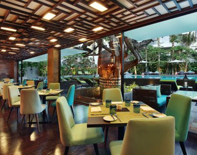 Bright, modern restaurant interior with green chairs and wooden tables set for dining. Large windows reveal a lush garden and outdoor seating area.
