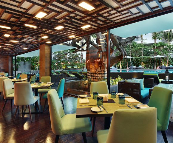 Bright, modern restaurant interior with green chairs and wooden tables set for dining. Large windows reveal a lush garden and outdoor seating area.