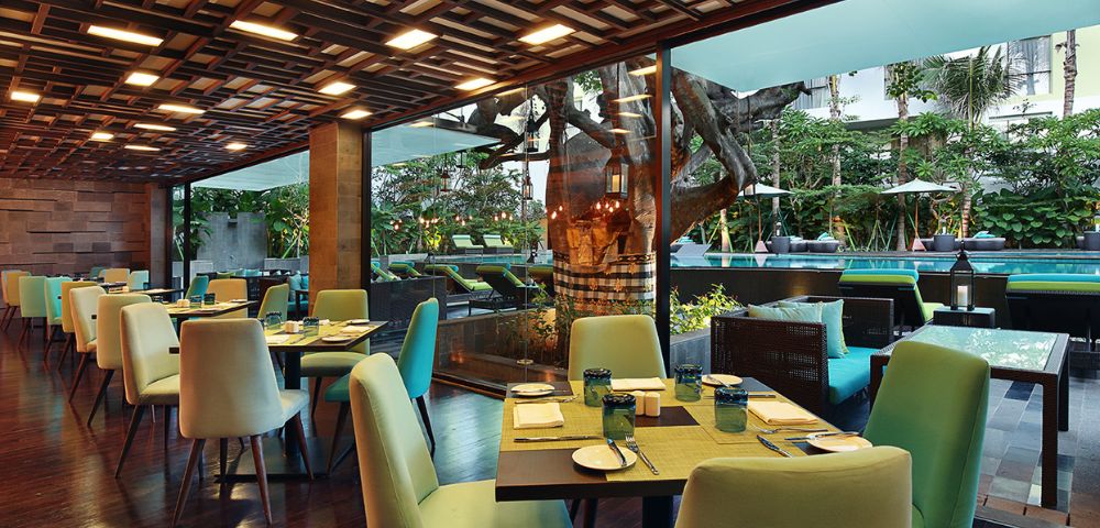 Bright, modern restaurant interior with green chairs and wooden tables set for dining. Large windows reveal a lush garden and outdoor seating area.
