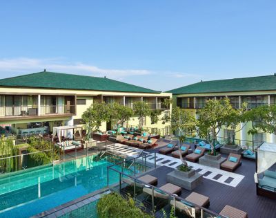 Luxurious resort courtyard featuring a tranquil pool, sun loungers, and greenery. Two-story buildings with green roofs under a clear blue sky.