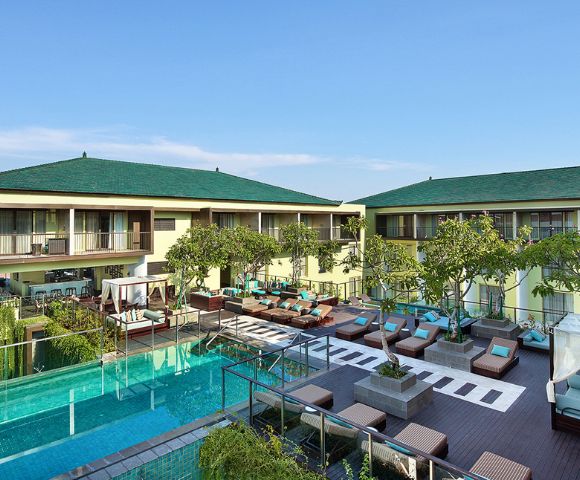Luxurious resort courtyard featuring a tranquil pool, sun loungers, and greenery. Two-story buildings with green roofs under a clear blue sky.