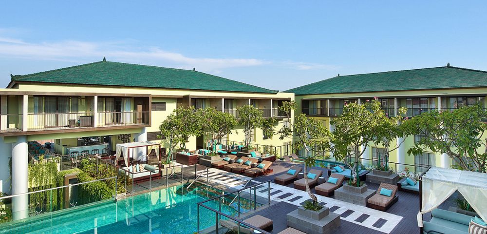 Luxurious resort courtyard featuring a tranquil pool, sun loungers, and greenery. Two-story buildings with green roofs under a clear blue sky.