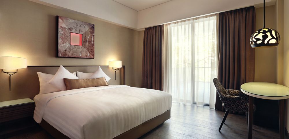 Modern hotel room with a large bed, white linens, and decorative pillows. Soft lighting and abstract art create a cozy, welcoming atmosphere.