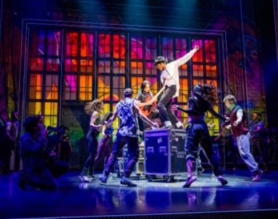 Energetic dance scene on stage with vibrant, multicolored backdrop. Dancers surround a performer on a platform, capturing excitement and movement.