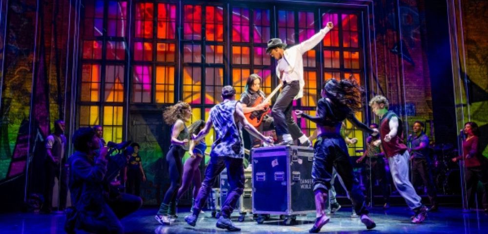 Energetic dance scene on stage with vibrant, multicolored backdrop. Dancers surround a performer on a platform, capturing excitement and movement.