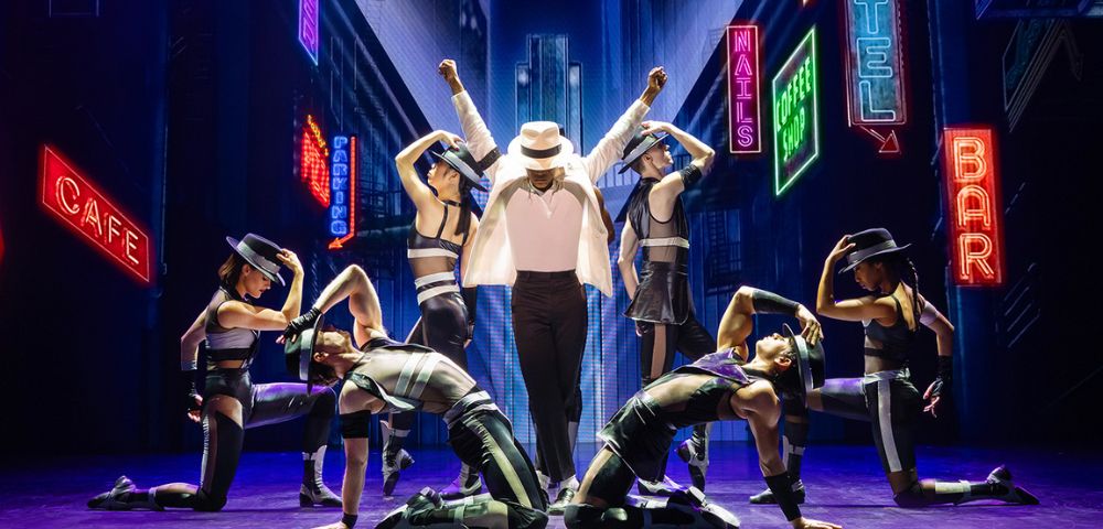 A performer in a white jacket and fedora stands center stage with six dancers posing around him. Neon city signs create a vibrant, energetic atmosphere.