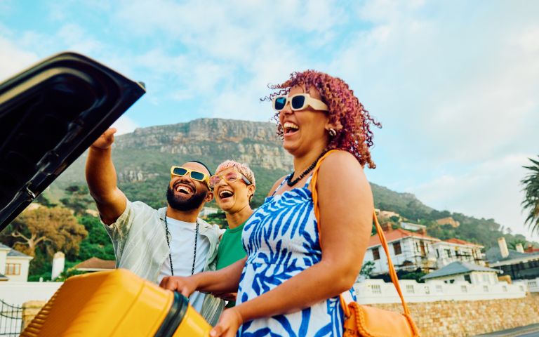 A woman in a blue and white dress holds a yellow suitcase while two men gesture, with a scenic mountain backdrop in a lively outdoor setting.