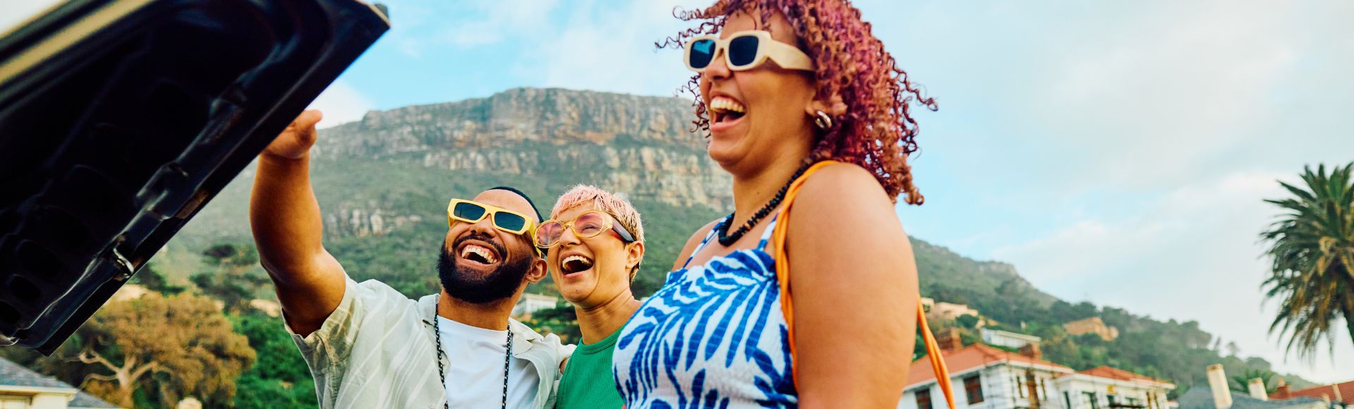 A woman in a blue and white dress holds a yellow suitcase while two men gesture, with a scenic mountain backdrop in a lively outdoor setting.