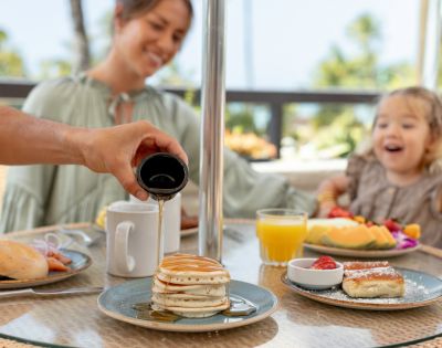 A person pours syrup on pancakes at an outdoor table with a joyful child and woman. Breakfast items and juice enhance the cheerful, sunny atmosphere.