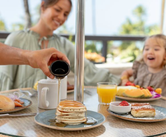A person pours syrup on pancakes at an outdoor table with a joyful child and woman. Breakfast items and juice enhance the cheerful, sunny atmosphere.