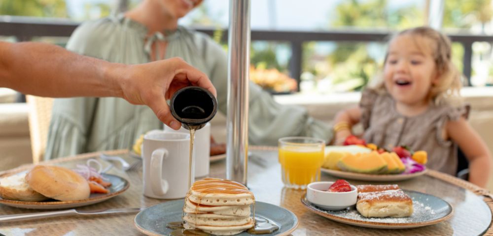 A person pours syrup on pancakes at an outdoor table with a joyful child and woman. Breakfast items and juice enhance the cheerful, sunny atmosphere.