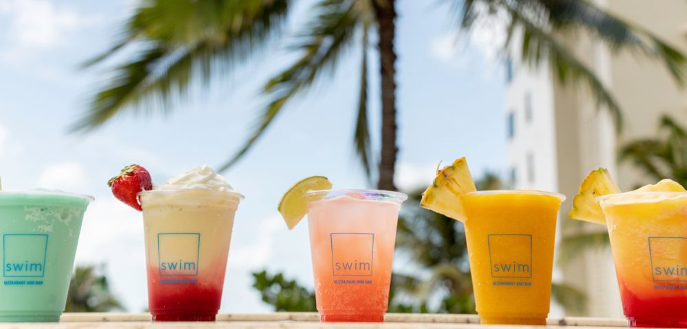 Tropical drinks on a wooden table, framed by a palm tree and blue sky. Each drink has unique garnishes, evoking a refreshing, beachy vibe.