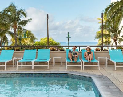 Two people relax on turquoise lounge chairs by a pool, surrounded by palm trees and a clear sky. The scene conveys a peaceful, tropical vacation vibe.