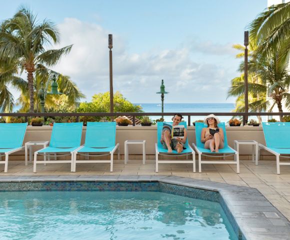 Two people relax on turquoise lounge chairs by a pool, surrounded by palm trees and a clear sky. The scene conveys a peaceful, tropical vacation vibe.