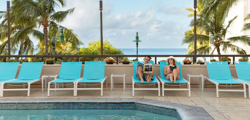 Two people relax on turquoise lounge chairs by a pool, surrounded by palm trees and a clear sky. The scene conveys a peaceful, tropical vacation vibe.