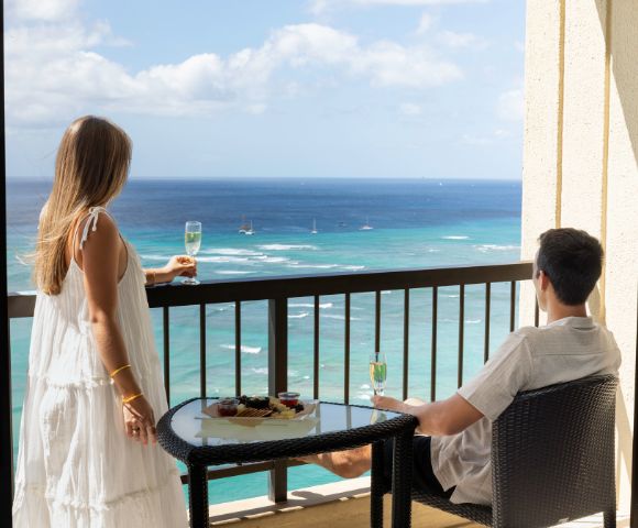 A couple on a balcony overlooks a calm ocean under a partly cloudy sky. They hold glasses of champagne next to a table with snacks, conveying relaxation.