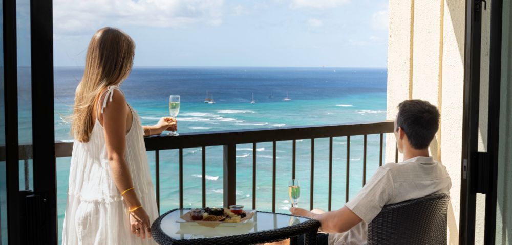 A couple on a balcony overlooks a calm ocean under a partly cloudy sky. They hold glasses of champagne next to a table with snacks, conveying relaxation.