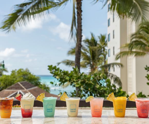 Tropical drinks in vibrant colors line a beachside counter with palm trees, blue sky, and ocean in the background; a relaxed, sunny vacation vibe.