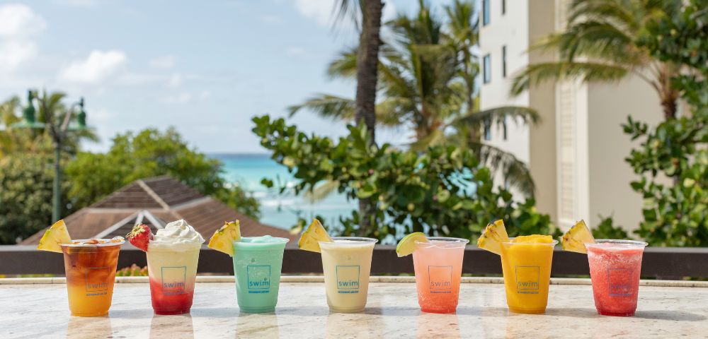 Tropical drinks in vibrant colors line a beachside counter with palm trees, blue sky, and ocean in the background; a relaxed, sunny vacation vibe.
