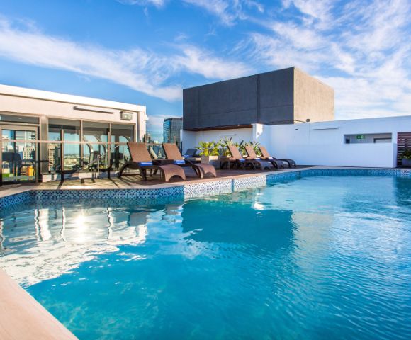 Rooftop pool with clear blue water and surrounding lounge chairs under a bright sky. Modern building backdrop, evoking a tranquil, luxurious vibe.