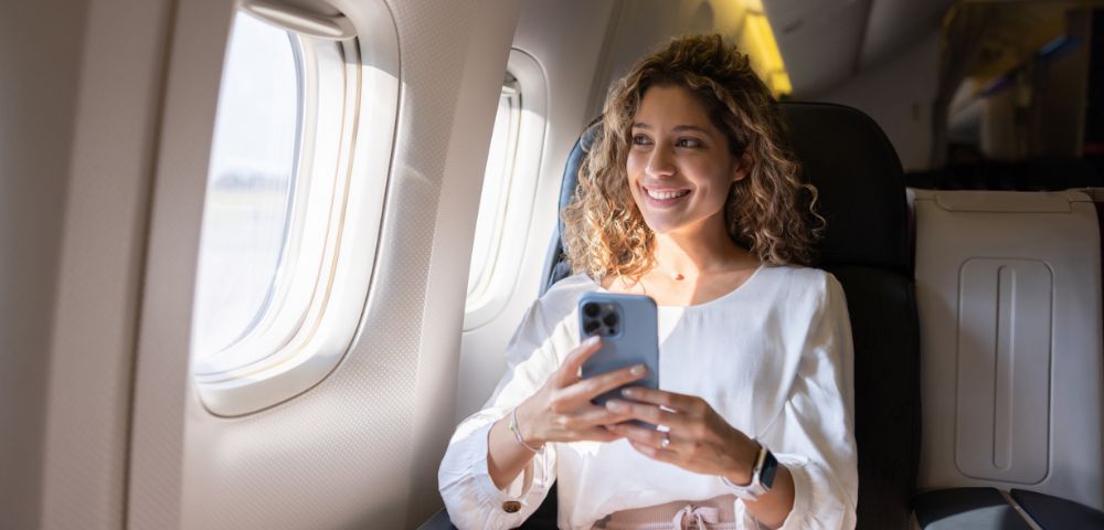 A woman with curly hair, wearing a white blouse and red belt, smiles while holding a smartphone on a plane. Sunlight streams through the window.