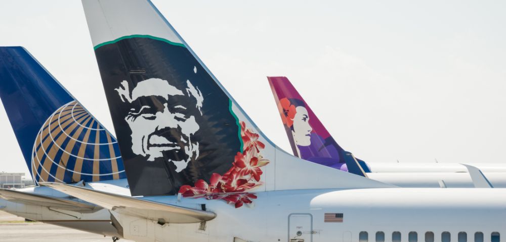 Three airplane tails are lined up at an airport, each showcasing distinct designs. The foreground tail features a monochrome face with red flowers, while the others display a colorful graphic and a globe logo. The sky is clear, giving a relaxed, airy feel.