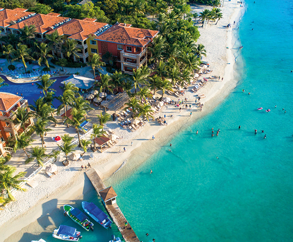 Aerial view of a vibrant tropical beach with turquoise water, lush palms, and colorful buildings. People relax on the sand, conveying a lively, cheerful atmosphere.