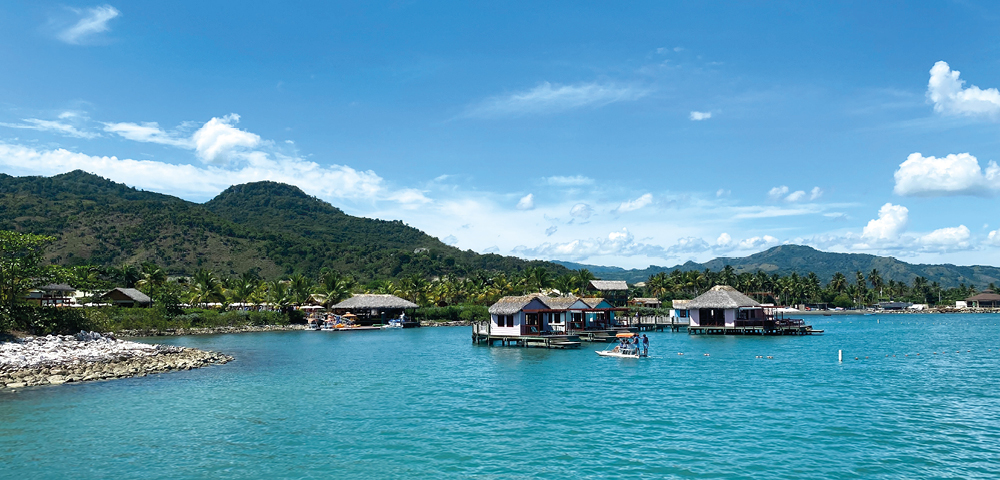 Scenic view of turquoise waters with stilted huts, lush green hills under a clear blue sky and fluffy clouds. A tranquil, idyllic tropical setting.