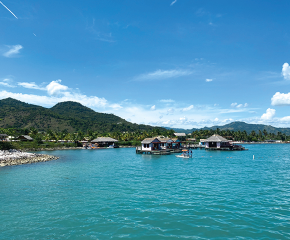 Scenic view of turquoise waters with stilted huts, lush green hills under a clear blue sky and fluffy clouds. A tranquil, idyllic tropical setting.