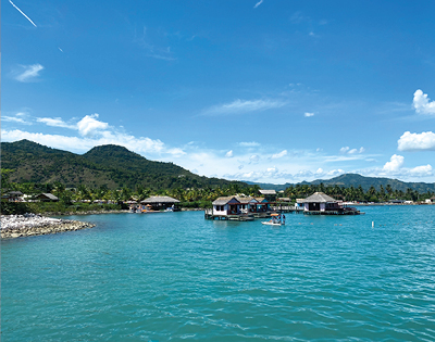 Scenic view of turquoise waters with stilted huts, lush green hills under a clear blue sky and fluffy clouds. A tranquil, idyllic tropical setting.