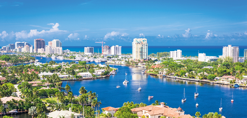 Aerial view of Fort Lauderdale, featuring a winding blue waterway lined with boats and bordered by lush greenery, urban buildings, and a bright, clear sky.